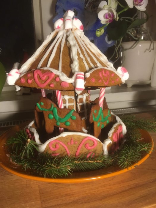 Handgjord pepparkaksmodell av en karusell dekorerad med glasyr och godisstångar på en tallrik.