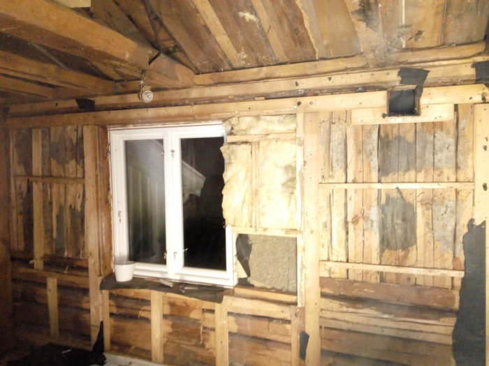 Delvis rivet timmervägg med synlig isolering och nytt fönster, stockstruktur och stöttande balkar synliga.