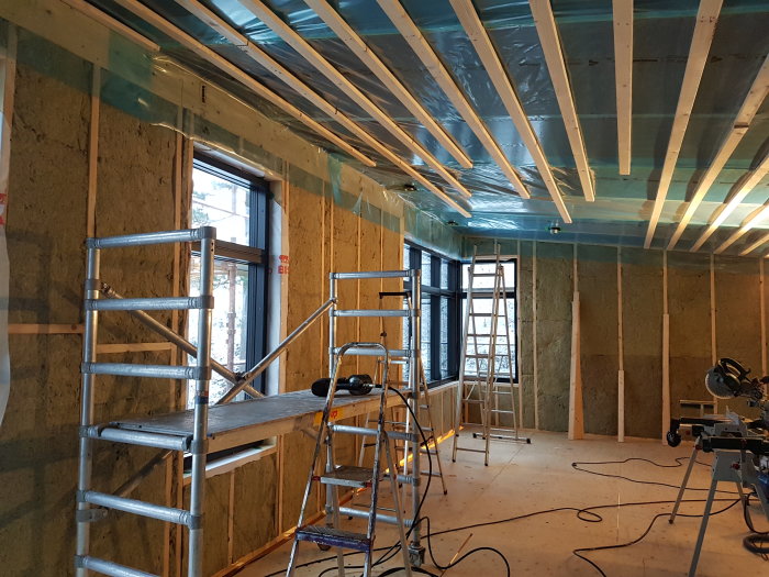 Renoveringsarbete med gles spikad på takbjälkar och isolering på väggar, samt en begagnad rullställning i rummet.