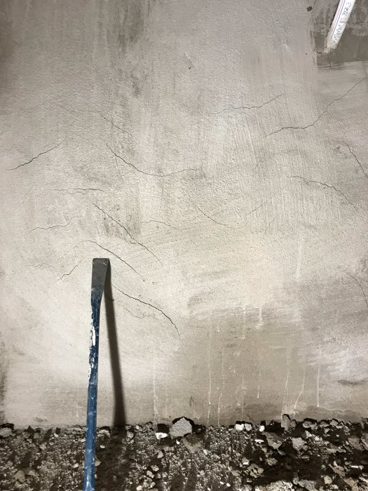 Nyputsad källarvägg med sprickor och avslagen puts nära golvet, en mejsel står lutad mot väggen.