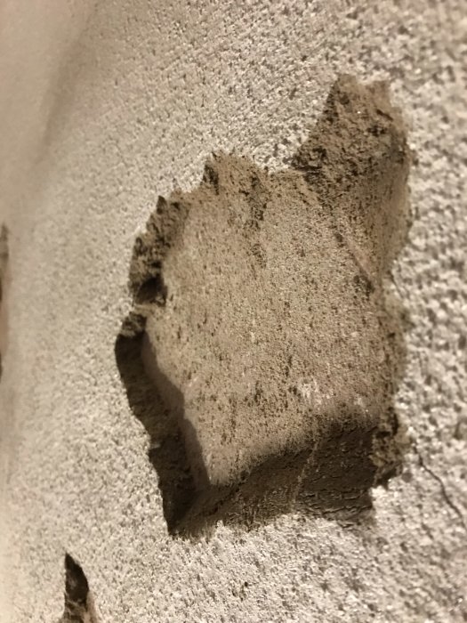 Närbild på en källarväggs hörn där putsen har fallit av, exponerande underliggande lager och sprickor.