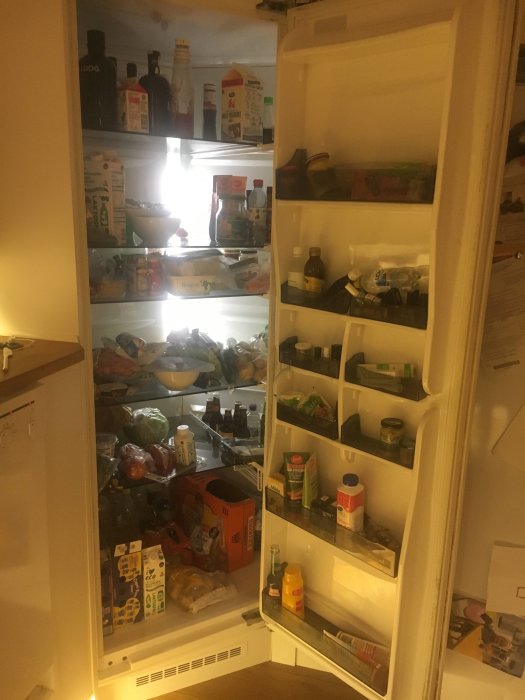 Öppet hörnkylskåp fullt med olika livsmedel och förpackningar, belyst inifrån.