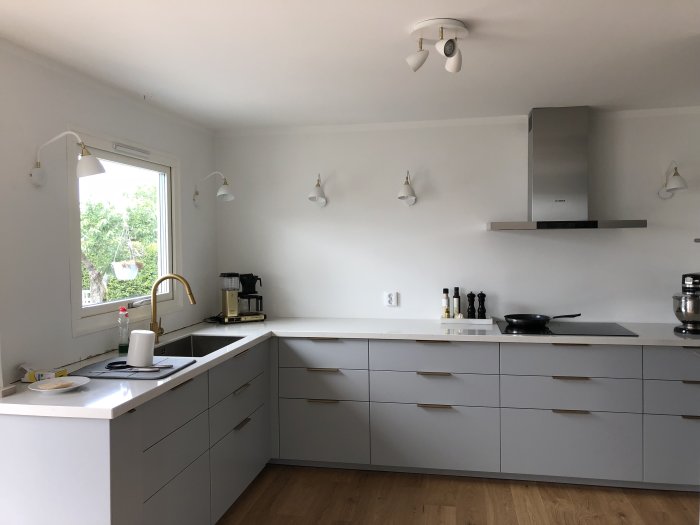 Ikea-kök utan hörnskåp med grå skåpfront, vitt kakel, guldig kran och rostfri fläkt.