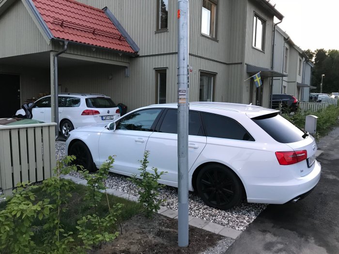 Delvis skymd vy av ett hus bakom en vit bil och en stolpe, med svensk flagga och grönska.