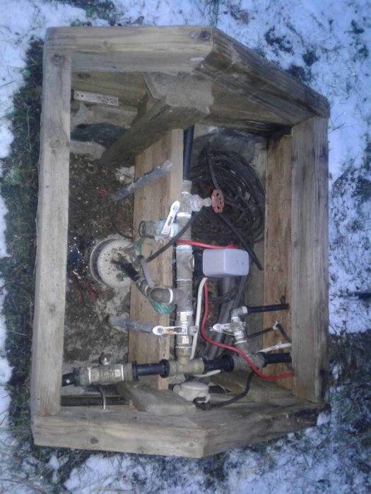 Vattenpumpsutrustning med tryckvakt och flera ledningar i ett brunnskydd, omgivet av snö.