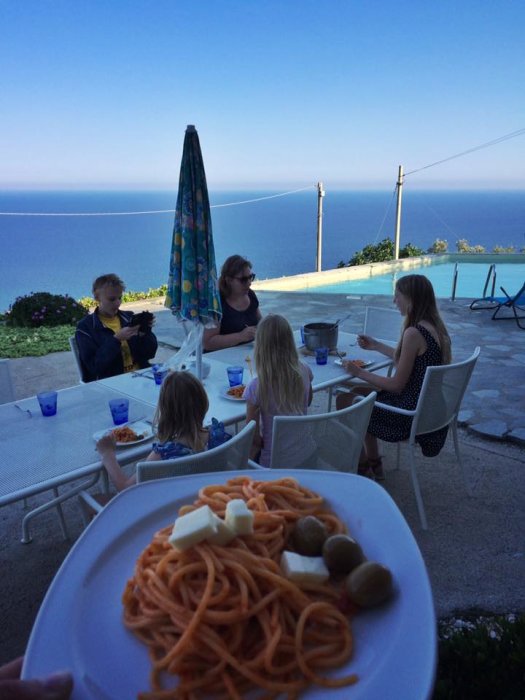 Familj äter måltid utomhus med utsikt över hav och pool, tallrik med pasta i förgrunden.