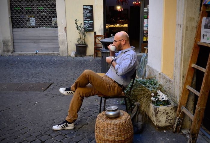 Man avnjuter kaffe utomhus vid ett café med gatuvy och rustik atmosfär.