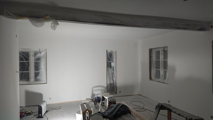 Vardagsrum under renovering med nymålade väggar och täckta fönster, renoveringsmaterial synligt.