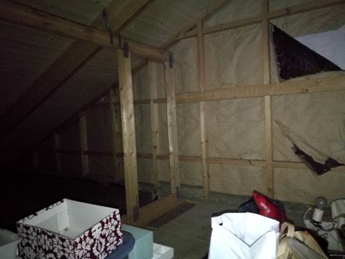 Otillräckligt belyst vind med synliga takstolar och isolermaterial, samt diverse föremål på golvet.