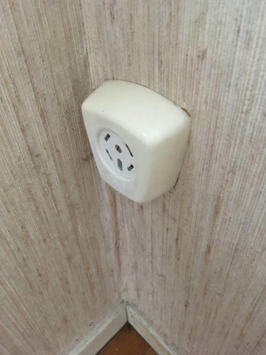 Gammalt beige telefonjack på en vägg med tapet, placerat vid golvlist i ett hörn.