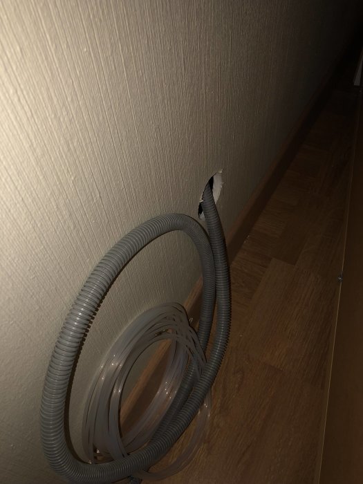 Flexibel skyddsslang med kabel utspridd på parkettgolv intill hål i tapetserad vägg.