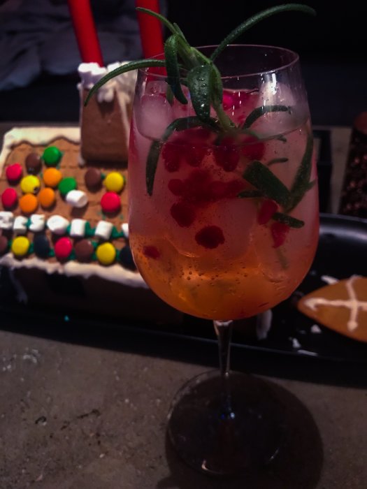 Rosaröd drink med is och gren av rosmarin framför en pepparkakshusverkstad.