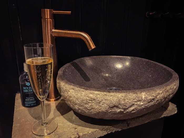 Handfat i sten med mässingskran och ett glas champagne, mot en svart vägg.