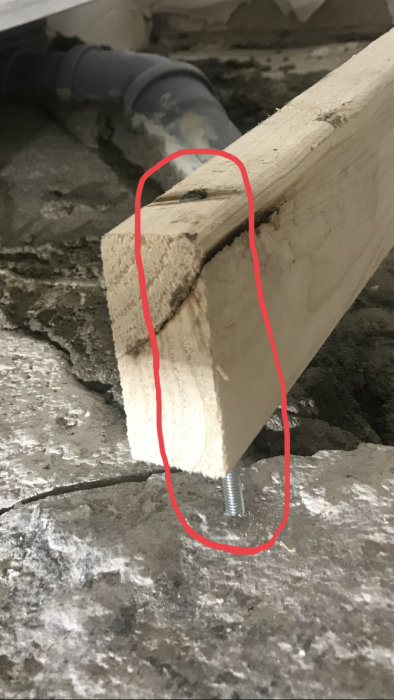En träregel med genomgående hål stödjer sig på en gängstång med bricka och mutter på en ojämn betongyta.