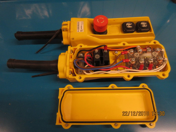 Två gula handkontroller för telfer öppnade med intern elektronik synlig, röd nödstopp och svarta upp/ned-knappar.