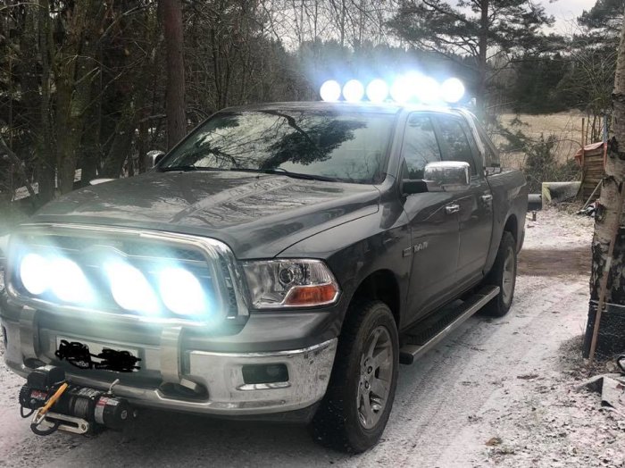 Pickup med extraljus på tak och front, parkerad på en grusväg med snöfläckar.