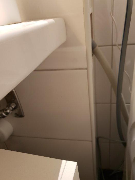 Delvis synligt handfat med vattenlåshål och bricka, vit kaklad vägg och rör i bakgrunden.