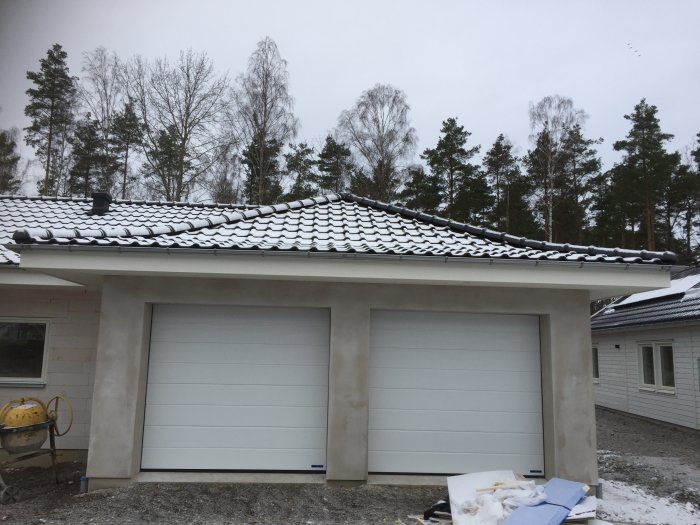 Två vita garageportar på ett hus med snötäckt tak, träd i bakgrunden och snö på marken.