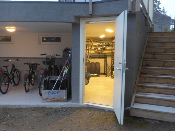 Ett öppet garage med cyklar och hyllor, trappor på höger sida och dörr öppen mot ett förråd.