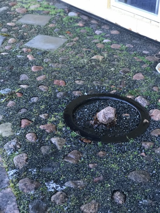Rund brunnslock i småstenbelagd mark med en liten spricka och växtlighet runt omkring.