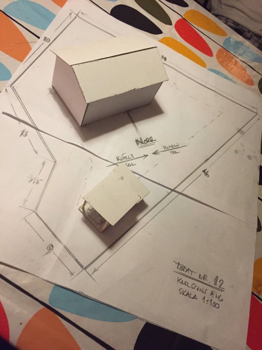 Modell av ett hus på ritad tomtplan med vägriktningar och skala 1:100 noterat.