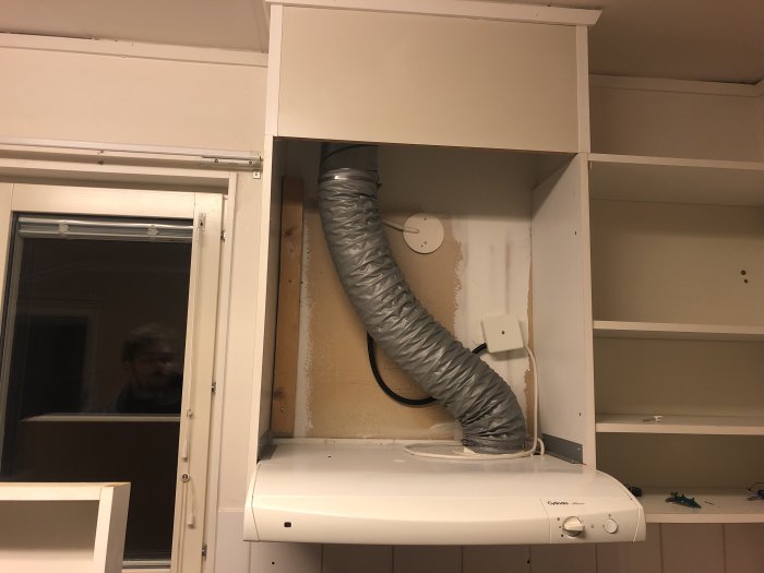 Köksskåp med öppen dörr, flexibel ventilationskanal och en spisfläkt, möjlig Alliancefläkt. Reflektion i fönster.