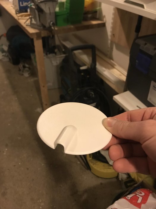 Hand håller ett vitt plastlock för elektrisk dosa i en rörig förrådsmiljö.