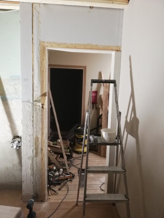 Dörröppning med ojämn betongvägg till vänster bredvid en dörrkarm, stegar och verktyg på golvet.