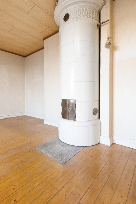 Gammaldags kakelugn i ett hörn av ett rum med trägolv och vitmålade väggar, indikativt för husets återställningspotential.
