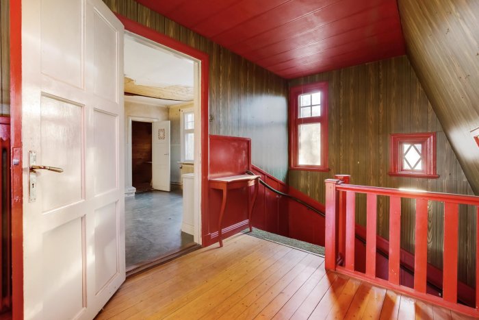 Interiör av äldre hus med röda och träfärgade panelväggar, trägolv och öppen dörr.