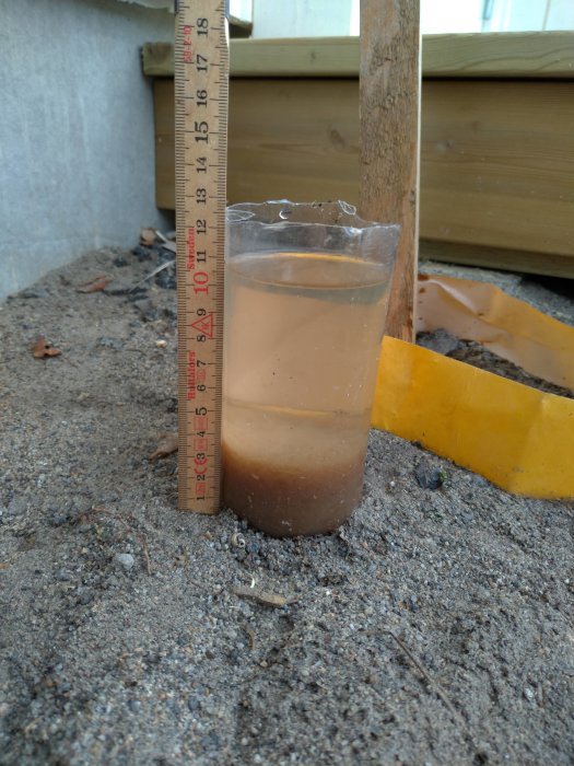 Ett genomskinligt måttglas fyllt med 50 cl brunaktigt vatten visar sedimentering, står på grusig mark bredvid en tumstock.
