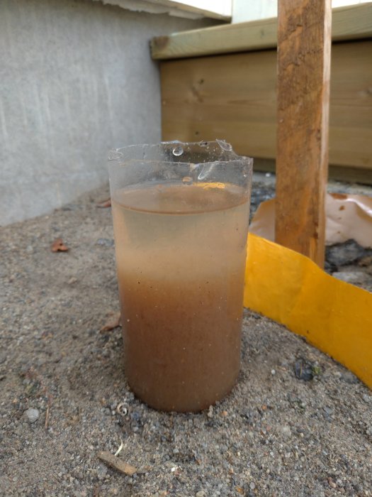 Mörkbrunt, grumligt vatten i en genomskinlig plastmugg med bakgrund av husets nock och avluftning.