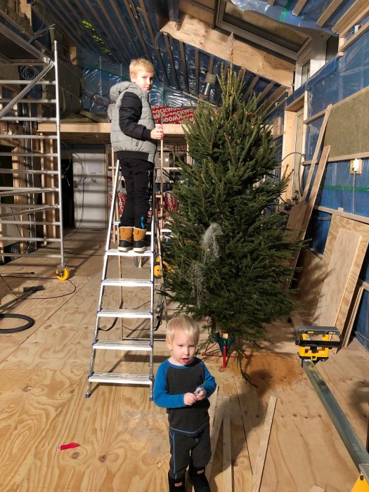 Två barn i ett under-renovering-hus med synlig konstruktion och en julgran.