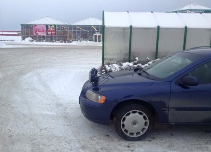 Blå bil med ovanligt monterade högtalare på motorhuven parkerad på snötäckt parkeringsplats.