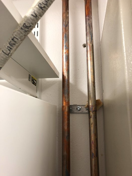 Missfärgade kopparrör med ojämn patina i ett pannrum, stödd mot vit vägg och elskåp.