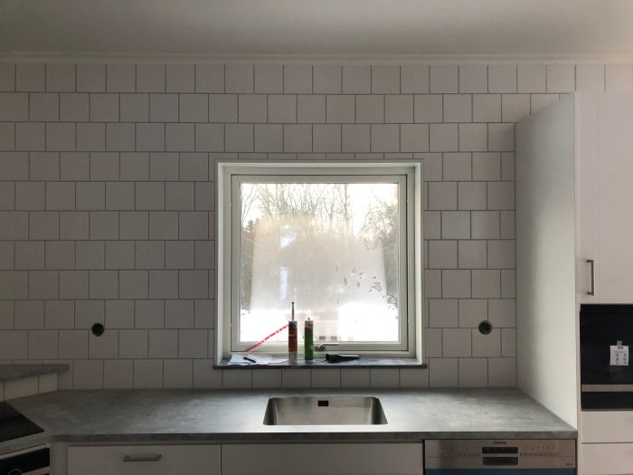 Kök med diskbänk ej centrerad under fönstret, vita kakelplattor på väggen och grå bänkskiva.