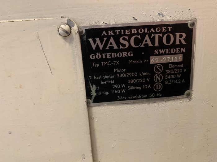 Informationsskylt på en äldre maskin från Wascator med tekniska specifikationer, monterad på en vit vägg.