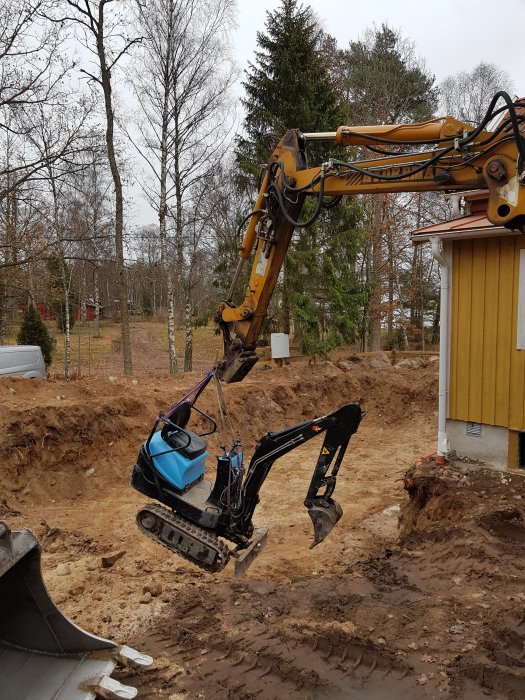 Minigrävare i arbete med schaktning av mark för garagegrund, omgiven av lövträd och ett gult hus.