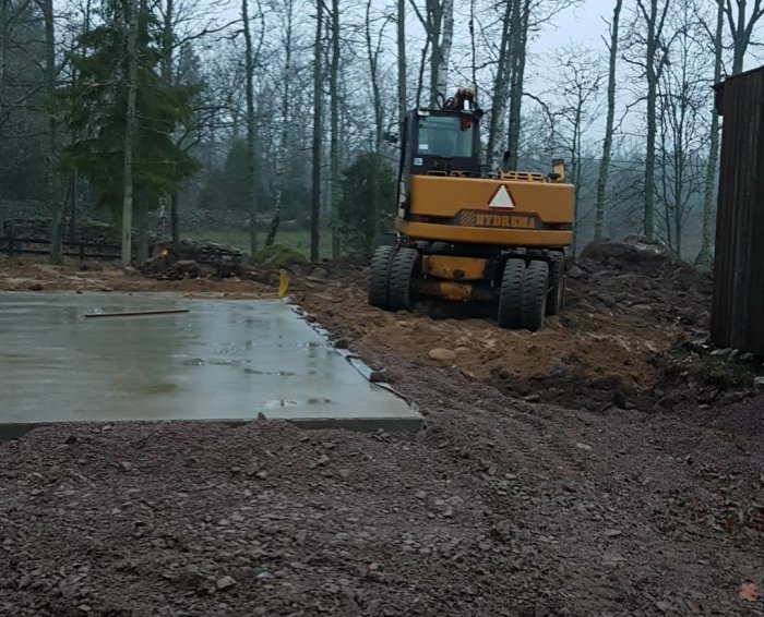 Grundläggning av garage med betongplatta och en gul grävmaskin på plats i skogsmiljö.