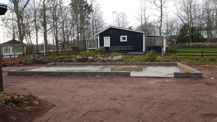 Nybyggd betonggrund för ett garage med markerade hörnstenar och en husbakgrund.