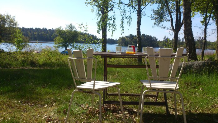 Utsikt över en sjö från en trädgård med ett bord och stolar, en karaff och glas i solsken.