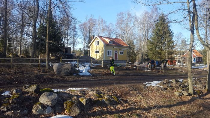 En gul villa med glasveranda omgivet av träd och snöfläckar, personer arbetar i trädgården.