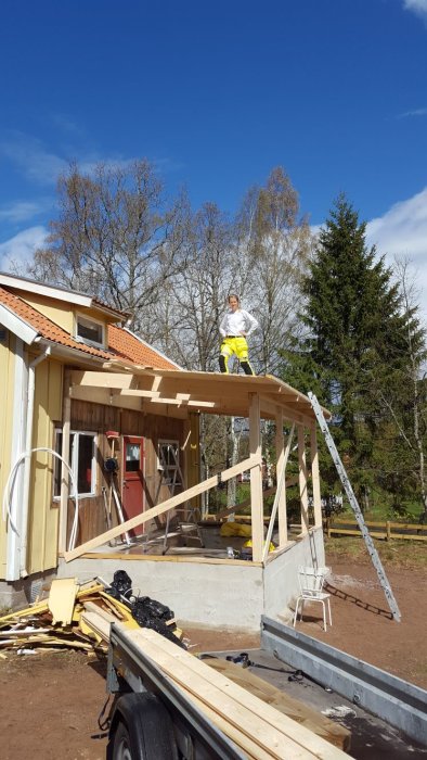 Person står på tak till glasveranda under renovering med träd i bakgrunden.