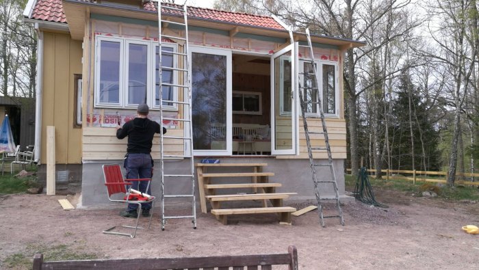 Renovering av glasveranda med person som arbetar, omgiven av stegar och verktyg, med skog i bakgrunden.