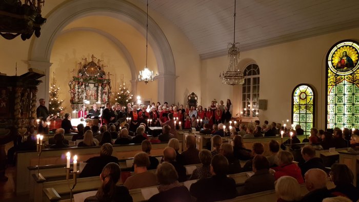 Kör framförar julmusik i kyrkan med åhörare som håller i levande ljus, omgiven av juldekorationer.