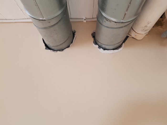 To ventilationsrør gennem en betongvæg med ujævne huller omkring. Rørene er sølvfarvede og omgivet af hvidt isoleringsmateriale.