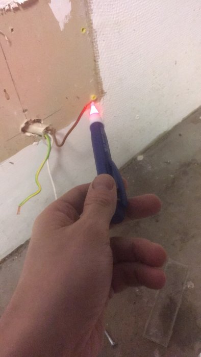 Hand håller en voltsticka som lyser rött mot en brun el-kabel i en vägg, med vit och gul/grön kabel synlig.