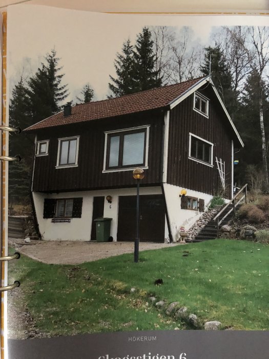 Tvåvåningshus med mörk träpanel och vita fönster mot en skogsbakgrund, med trappa och garage.