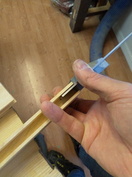 En hand som håller en träbit med tappar bredvid ett tapphål, verktyg och träspån syns.