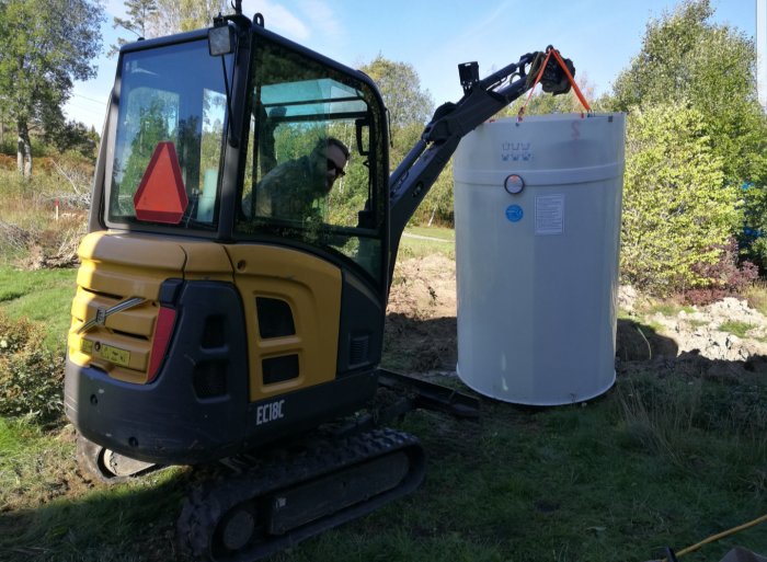 Minigrävare som lyfter en stor vit cistern utomhus på ett byggarbetsområde.